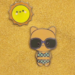 May include: A brown cat wearing sunglasses and a green and blue swimsuit with hearts. The cat is standing on a yellow sandy surface with a yellow sun with a smiling face above it.