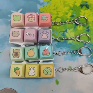 May include: A collection of colorful keycaps with various cute illustrations, including a frog, mushroom, snail, bear, orange, and watermelon. Each keycap is attached to a keychain, offering a fun and unique accessory.