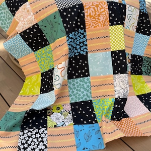 May include: A colorful patchwork quilt with a variety of patterned squares. The quilt features squares of black with white stars, floral prints, and geometric designs in shades of blue, green, orange, and white. The quilt is draped over a surface.