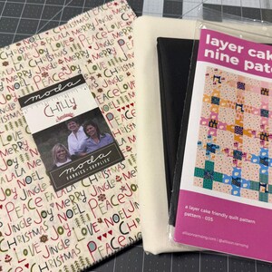 May include: A stack of fabric squares featuring a cream background with repeating red, green, and black holiday-themed text and icons, alongside a black fabric bundle and a quilt pattern booklet titled Layer Cake Nine Patch, pattern 035.