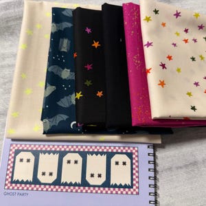 May include: A stack of six folded fabric pieces featuring various patterns including bats, multicolored stars, and solid colors, resting on a spiral-bound notebook with a graphic of five white ghosts and the text Ghost Party printed on the cover.