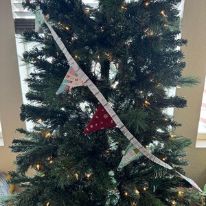 May include: A decorated Christmas tree with a garland of small, colorful pennant flags. The flags are red, green, and blue with festive patterns. The tree is lit with small, warm white lights.