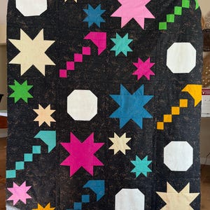 May include: A rectangular quilt featuring a black background with a speckled texture. The design includes various geometric shapes in bright colors, including pink, teal, yellow, and white stars, octagons, and pixelated arrow patterns.