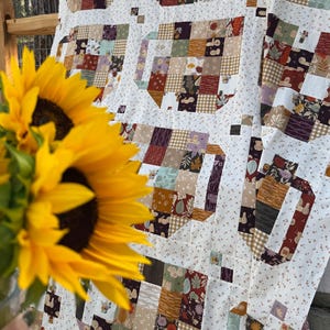 May include: A patchwork quilt featuring a repeating teapot motif constructed from various fabric squares in shades of brown, orange, green, and cream, set against a white background with a small floral print, partially obscured by bright yellow sunflowers.