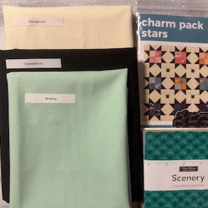 May include: A collection of quilting supplies including three folded fabric pieces in cream, black, and mint green, each labeled with white paper tags. Beside them is a charm pack stars pattern booklet and a stack of Scenery fabric squares.