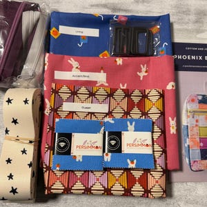 May include: A sewing kit for a Phoenix Backpack including blue fabric with small orange motifs, pink fabric with white rabbit patterns, a geometric print fabric, two blue fabric squares, a roll of cream webbing with black stars, and a pattern booklet.