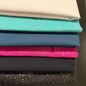 May include: A stack of fabric samples in various colors. The top layer is white, followed by teal with gold speckles, navy blue, hot pink, black, and a black fabric with gold speckles. The fabrics are neatly folded.
