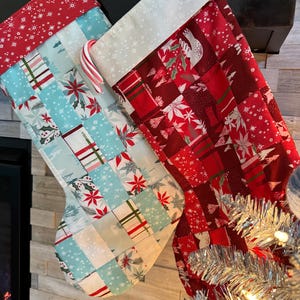 May include: Two Christmas stockings with patchwork designs. One is light blue with red and white accents, the other is red with white accents. A candy cane is placed in the light blue stocking. Both stockings have white cuffs and red hanging loops.