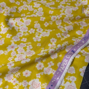 May include: Yellow fabric with a white and pink floral pattern. The fabric has a repeating design of cherry blossoms. A purple ribbon with the text "PAPER CUTS 2" and "SOCIETY" is visible.