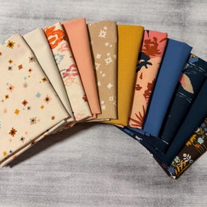 May include: A collection of folded fabric squares in various colors and patterns. The fabrics include floral, geometric, and solid designs in shades of cream, peach, mustard, and navy. Ideal for quilting or crafting projects.