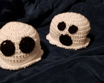 May include: Two handmade crochet ghost hats. Each white hat features large black eyes and a mouth, giving them a spooky appearance. The hats are displayed on a dark blue, textured surface.