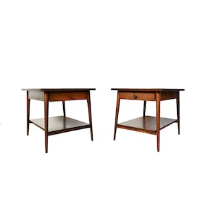 May include: Two matching mid-century modern end tables with a single drawer and a lower shelf. The tables are made of dark wood and have a simple, elegant design.