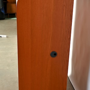 May include: A tall, wooden cabinet with a curved top and a reddish-brown finish. The cabinet has two black plastic grommets on the side.