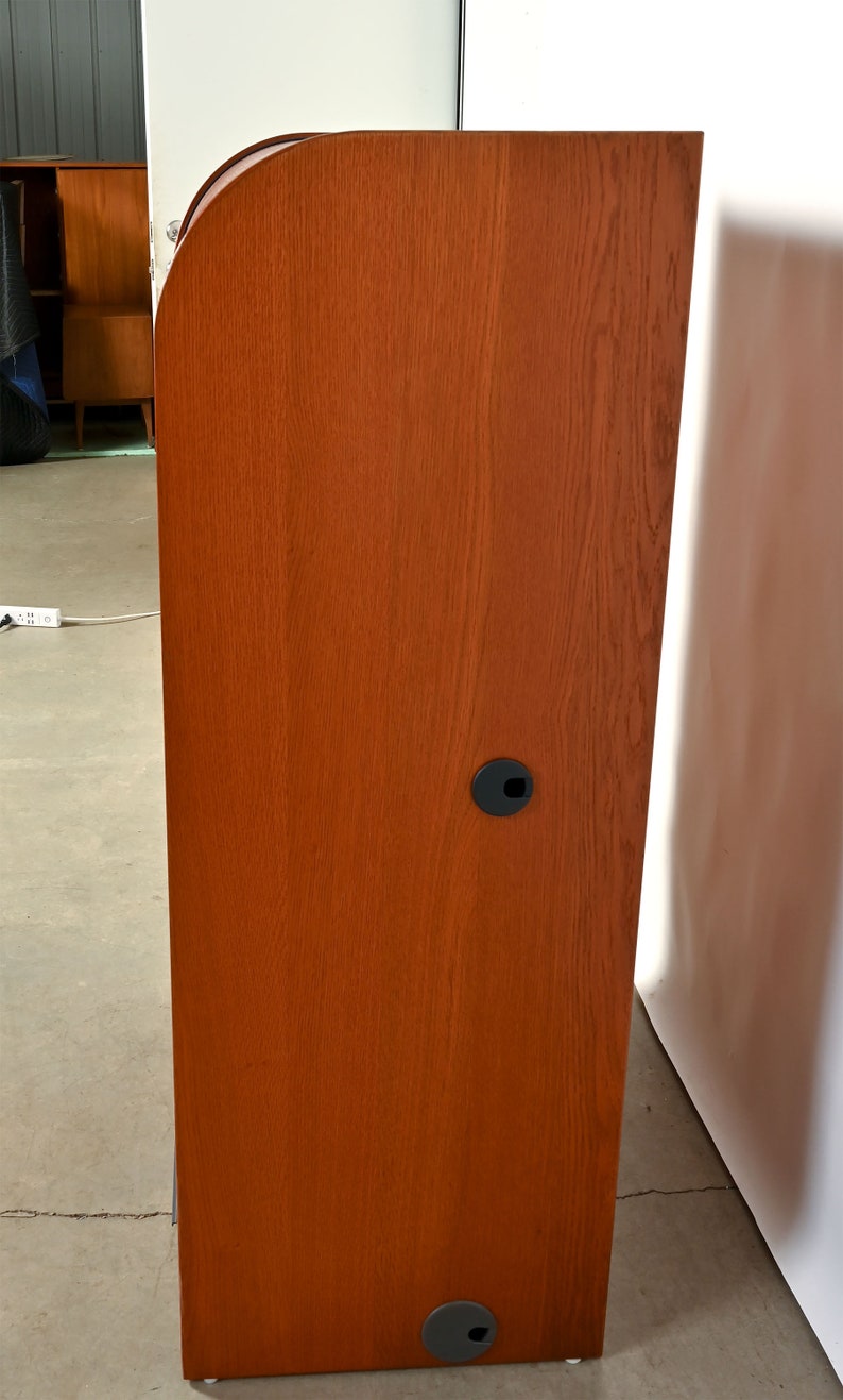 May include: A tall, wooden cabinet with a curved top and a reddish-brown finish. The cabinet has two black plastic grommets on the side.