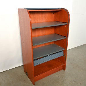 May include: A wooden cabinet with a curved front and a sliding door. The cabinet has four shelves and a drawer. The shelves are gray and the cabinet is a reddish-brown color.