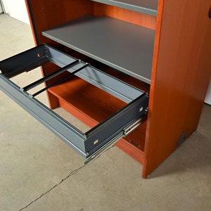 May include: A wooden cabinet with a gray metal drawer that is pulled out. The drawer has a metal frame and is on metal runners. The cabinet is a reddish-brown color.