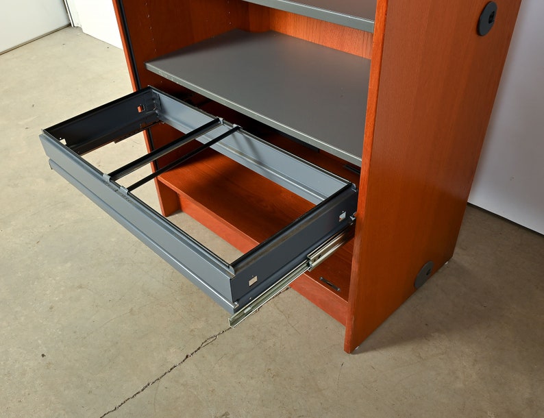 May include: A wooden cabinet with a gray metal drawer that is pulled out. The drawer has a metal frame and is on metal runners. The cabinet is a reddish-brown color.