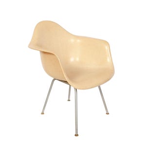May include: A vintage, light beige armchair with a curved seat and armrests. The chair has a textured surface and is supported by four slender, silver-colored metal legs. The legs have small, round, brown feet.