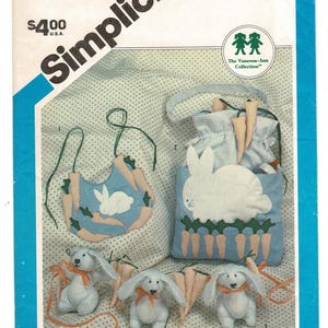 May include: A Simplicity 6427 sewing pattern envelope featuring a blue bib with a white rabbit and carrots, a blue drawstring bag with a white rabbit and carrot applique, and three small blue stuffed rabbits with orange ribbons and carrot accents.