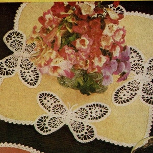 May include: A yellow placemat with a scalloped edge and three white crocheted butterfly accents. A bouquet of pink and red flowers sits on the placemat. The placemat is on a dark surface.
