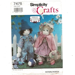 May include: A Simplicity Crafts sewing pattern envelope 7476 featuring two fabric dolls. One doll wears blue overalls and a blue knit hat, while the other wears a pink cardigan, a floral dress, and a floral bonnet. The text includes Shirley Botsford Designs.