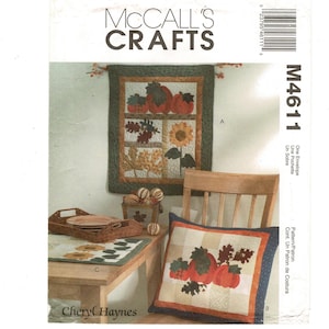 May include: A McCall's Crafts pattern envelope featuring autumn-themed quilted projects. The image showcases a wall hanging, pillow, and table runner with pumpkin and leaf designs. The color palette includes orange, green, and beige, evoking a fall harvest theme.