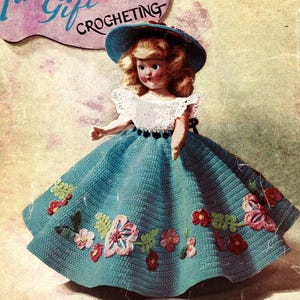 Peut inclure: Une robe de poupée en crochet avec une jupe bleue et un corsage blanc. La jupe est ornée d'un motif floral avec des fleurs roses, rouges et vertes. La poupée porte un chapeau bleu.