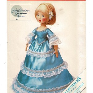 May include: A porcelain doll in a light blue satin gown with white lace trim. The doll has blonde hair, rosy cheeks, and red lips. The doll's dress has multiple layers of lace and a fitted bodice. The doll is part of the "Soft Porcelain Creations Janet" collection.