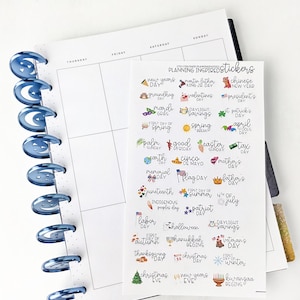 May include: A planner with blue spiral binding and a sheet of holiday-themed stickers. The stickers include text and small illustrations for various holidays and events, such as New Year's Day, Valentine's Day, and Christmas Eve.