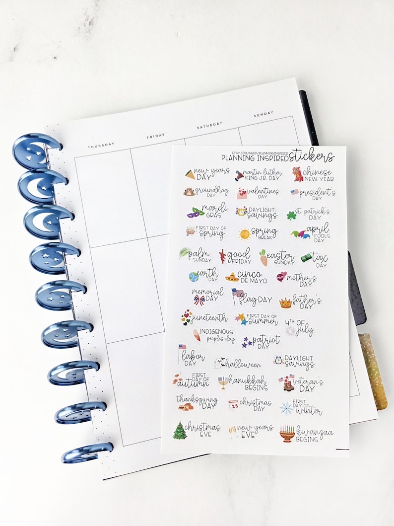 May include: A planner with blue spiral binding and a sheet of holiday-themed stickers. The stickers include text and small illustrations for various holidays and events, such as New Year's Day, Valentine's Day, and Christmas Eve.