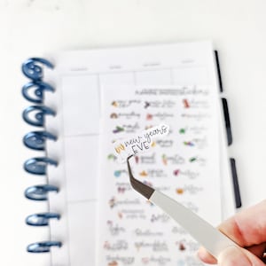May include: A planner sticker with the text "new years EVE" is held by tweezers. The sticker is white with black lettering and a gold champagne glass graphic. The sticker is on a sheet of planner stickers with various designs.