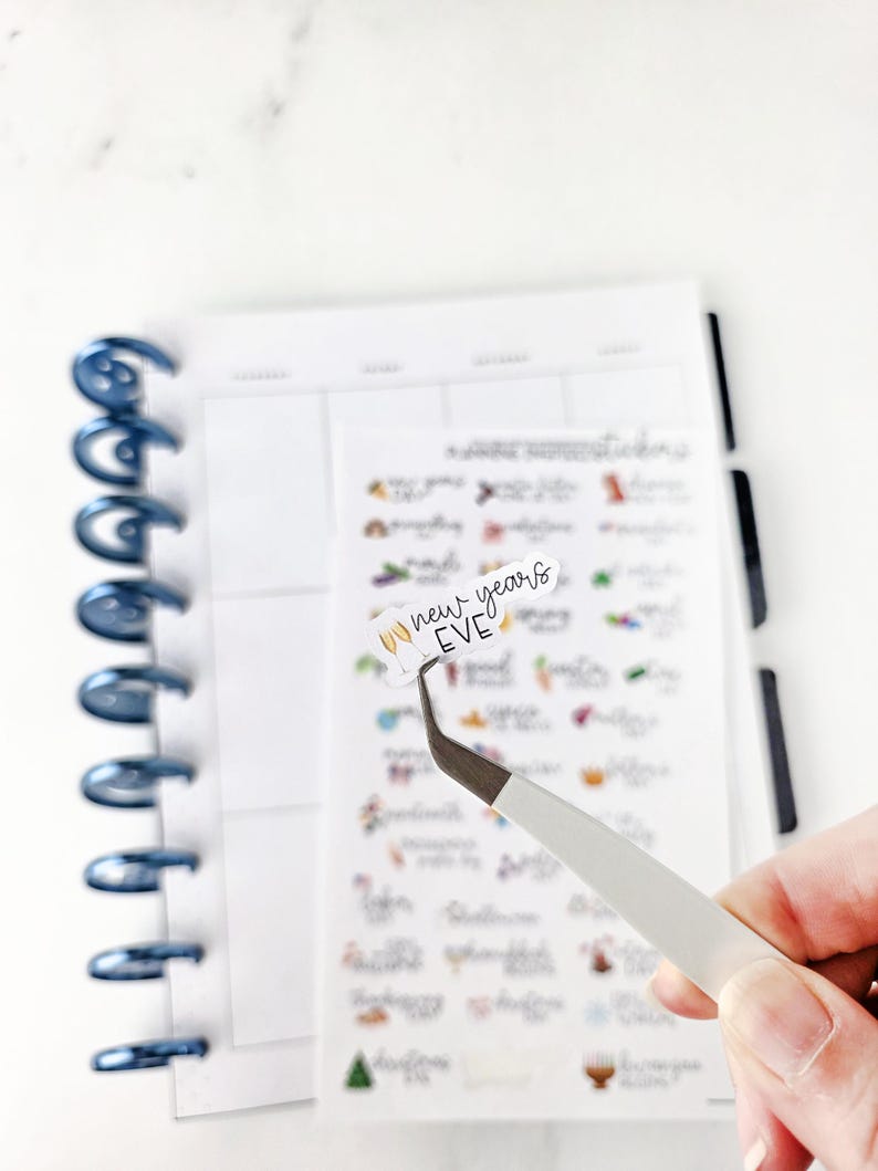 May include: A planner sticker with the text "new years EVE" is held by tweezers. The sticker is white with black lettering and a gold champagne glass graphic. The sticker is on a sheet of planner stickers with various designs.