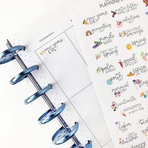 May include: A planner with a blue metal binding, open to a Thursday page with "New Year's Eve" written on it. A sheet of holiday-themed stickers is visible, with text like "New Year's Day," "Valentine's," and "Labor Day."