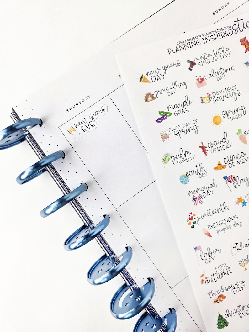 May include: A planner with a blue metal binding, open to a Thursday page with "New Year's Eve" written on it. A sheet of holiday-themed stickers is visible, with text like "New Year's Day," "Valentine's," and "Labor Day."