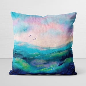 May include: Square throw pillow with a landscape painting. The artwork features a blue and green ocean scene with a pink and blue sky. Three birds are flying in the sky. The pillow has a textured appearance.