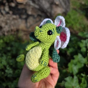 K&ouml;nnte beinhalten: Ein kleines H&auml;kel-Drachenspielzeug in leuchtendem Gr&uuml;n mit einem hellgelben Bauch, dunkelgr&uuml;nen Fl&uuml;geln und einer gro&szlig;en wei&szlig;en Blume mit rosa Mitte am Kopf. Die Figur hat schwarze Sicherheitsaugen aus Kunststoff.