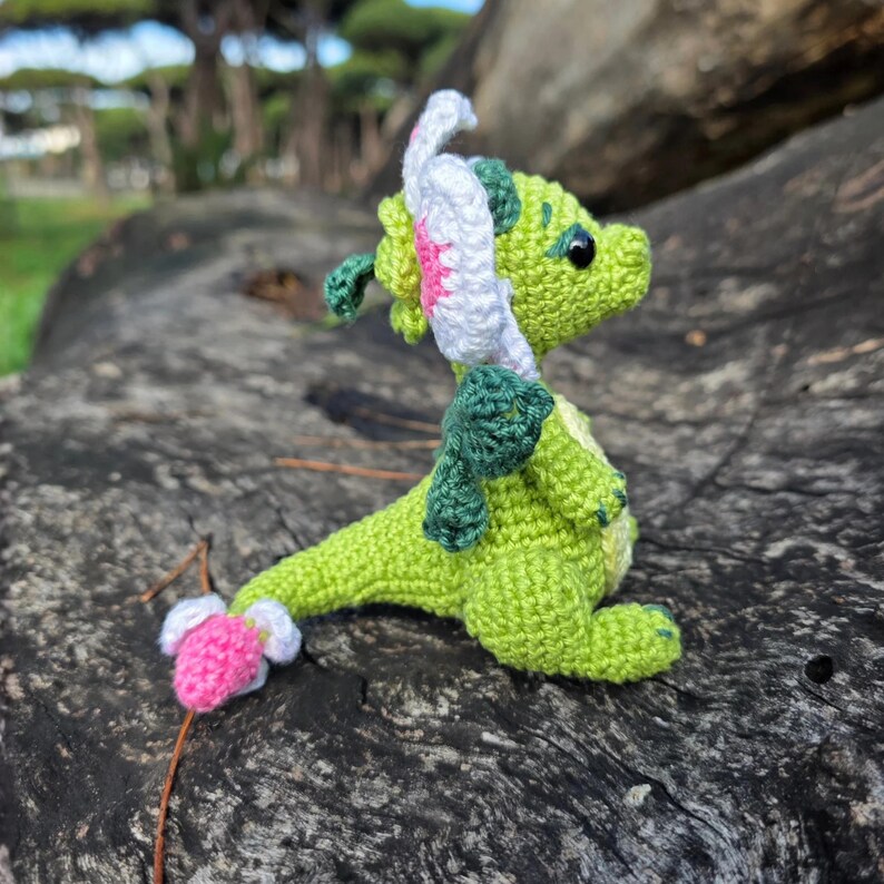 K&ouml;nnte beinhalten: Ein kleines, geh&auml;keltes gr&uuml;nes Drachen-Stofftier mit dunkelgr&uuml;nen Fl&uuml;geln, einem wei&szlig;-rosa bl&uuml;tenf&ouml;rmigen Kopfschmuck und einer rosa-wei&szlig;en Schwanzspitze. Die Figur hat schwarze Perlenaugen und eine strukturierte Garnoberfl&auml;che.