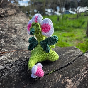 K&ouml;nnte beinhalten: Eine geh&auml;kelte Kreatur mit hellgr&uuml;nem K&ouml;rper, dunkelgr&uuml;nen Fl&uuml;geln und einem gro&szlig;en blumenf&ouml;rmigen Kopf mit wei&szlig;en Bl&uuml;tenbl&auml;ttern und einer leuchtend rosa Mitte. Der Schwanz endet in einer kleinen wei&szlig;-rosa Bl&uuml;tenknospe und hat eine gr&uuml;ne Schnauze.