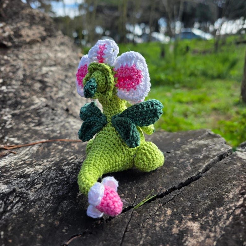 K&ouml;nnte beinhalten: Eine geh&auml;kelte Kreatur mit hellgr&uuml;nem K&ouml;rper, dunkelgr&uuml;nen Fl&uuml;geln und einem gro&szlig;en blumenf&ouml;rmigen Kopf mit wei&szlig;en Bl&uuml;tenbl&auml;ttern und einer leuchtend rosa Mitte. Der Schwanz endet in einer kleinen wei&szlig;-rosa Bl&uuml;tenknospe und hat eine gr&uuml;ne Schnauze.