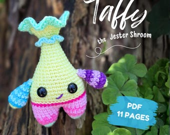 May include: A crochet plush toy shaped like a mushroom with a pale yellow body, a ruffled light green cap, and pink legs. It features black safety eyes, a small embroidered smile, and multicolored arms. Text reads: Taffy the Jester Shroom, PDF 11 PAGES, English (US).