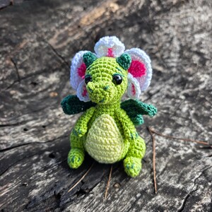 K&ouml;nnte beinhalten: Eine kleine, geh&auml;kelte gr&uuml;ne Drachenfigur mit hellgelbem Bauch, dunkelgr&uuml;nen Fl&uuml;geln und einem gro&szlig;en, bl&uuml;tenf&ouml;rmigen Kopfschmuck in Wei&szlig; und Pink. Das Wesen hat schwarze Perlenaugen und ist mit detaillierten Maschen gefertigt.