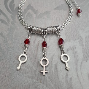 Könnte beinhalten: Ein silbernes Kettenarmband mit drei baumelnden Anhängern. Jeder Anhänger ist mit einer roten Kristallperle und einem silbernen Symbol verziert. Die Symbole sind ein Kreis mit einem nach unten zeigenden Pfeil, ein Kreis mit einem Kreuz und ein Kreis mit einem nach unten zeigenden Pfeil.