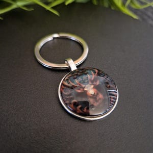 May include: A round silver keychain with a detailed image of a deer's head. The deer is depicted in shades of brown, black, and red, with prominent antlers. The keychain has a shiny, metallic finish and a sturdy ring.