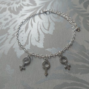 Op de afbeelding: Een zilveren enkelketting met drie hangende bedels in de vorm van de mannelijke en vrouwelijke geslachtsymbolen.