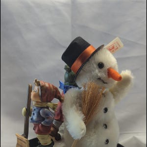 May include: A white plush snowman wearing a black top hat with an orange ribbon and holding a brown broom. A porcelain figurine of a child with a red hat and blue coat is holding a shovel next to the snowman. Both figures are on a black base.