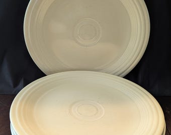May include: A stack of four cream-colored ceramic plates, with a larger plate on top. Each plate has concentric rings and a central indentation. The plates are arranged on a dark surface, with a black background.
