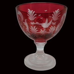 May include: A red glass goblet with a white etched design of birds and plants. The goblet has a clear glass stem and base.