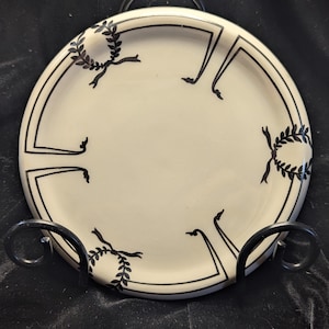 May include: A white ceramic plate with a black and white design of stylized leaves and bows around the edge. The plate is on a black stand.