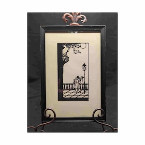 May include: A framed silhouette art piece depicting a person playing a musical instrument on a balcony, with a street lamp and tree in the background. The black and white artwork is set against a cream-colored background, framed in black, and displayed on a decorative metal stand.