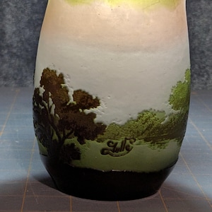 May include: A cylindrical glass vase with a landscape design. The vase features a gradient of colors, transitioning from green to white and peach. The base is black, and the rim is yellow-green. The landscape design includes trees and water.